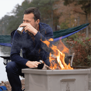 A man sips from a metal mug while sitting next to a Hot Ash Rambler Smokeless Fire Pit with a warm fire burning, set by a lake with a hammock in the background.