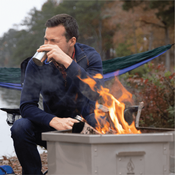 A man sips from a metal mug while sitting next to a Hot Ash Rambler Smokeless Fire Pit with a warm fire burning, set by a lake with a hammock in the background.
