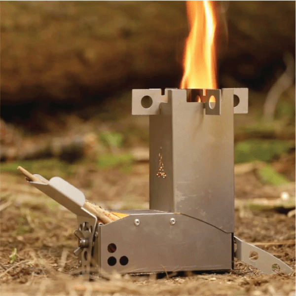 A Hot Ash rocket stove with a tall, intense flame burning from the top, fueled by small sticks in the feeder, set up outdoors on the forest floor.