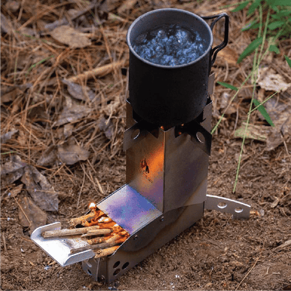A stainless steel Hot Ash rocket stove with a black camping pot on top, boiling water vigorously, fueled by small sticks in the side feeder, set in a natural outdoor environment.
