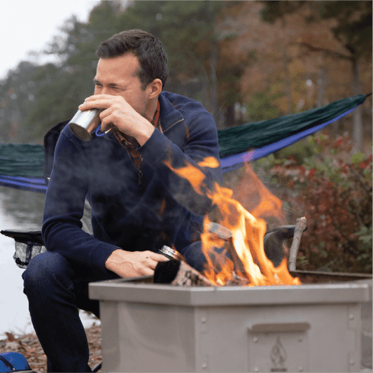 A man sips from a metal mug while sitting next to a Hot Ash Rambler Smokeless Fire Pit with a warm fire burning, set by a lake with a hammock in the background.